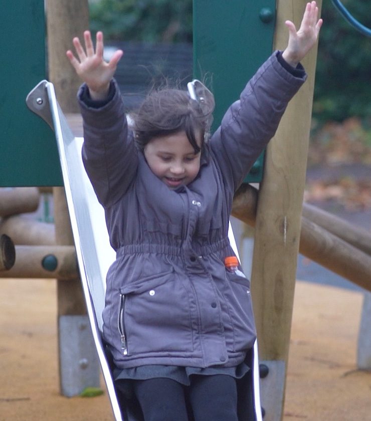 little girl going down a slide in a playground with arms in the air and dressed in a winter coat