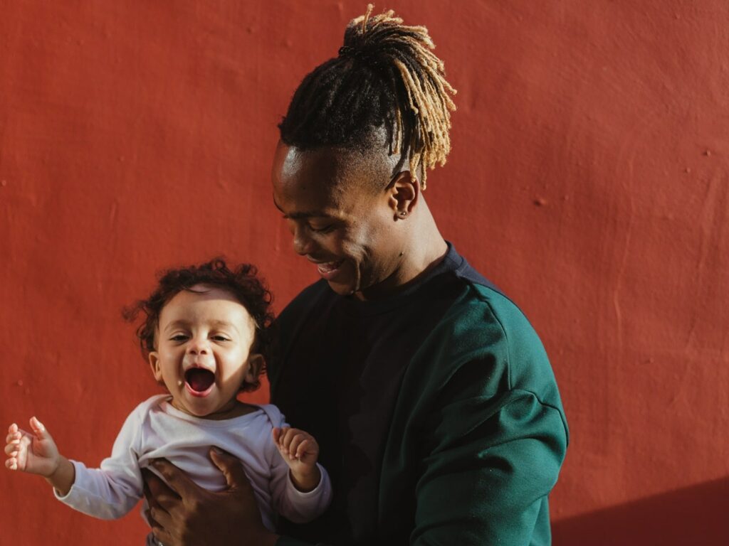 dad with dreadlocks standing in front of an orange wall with smiling baby