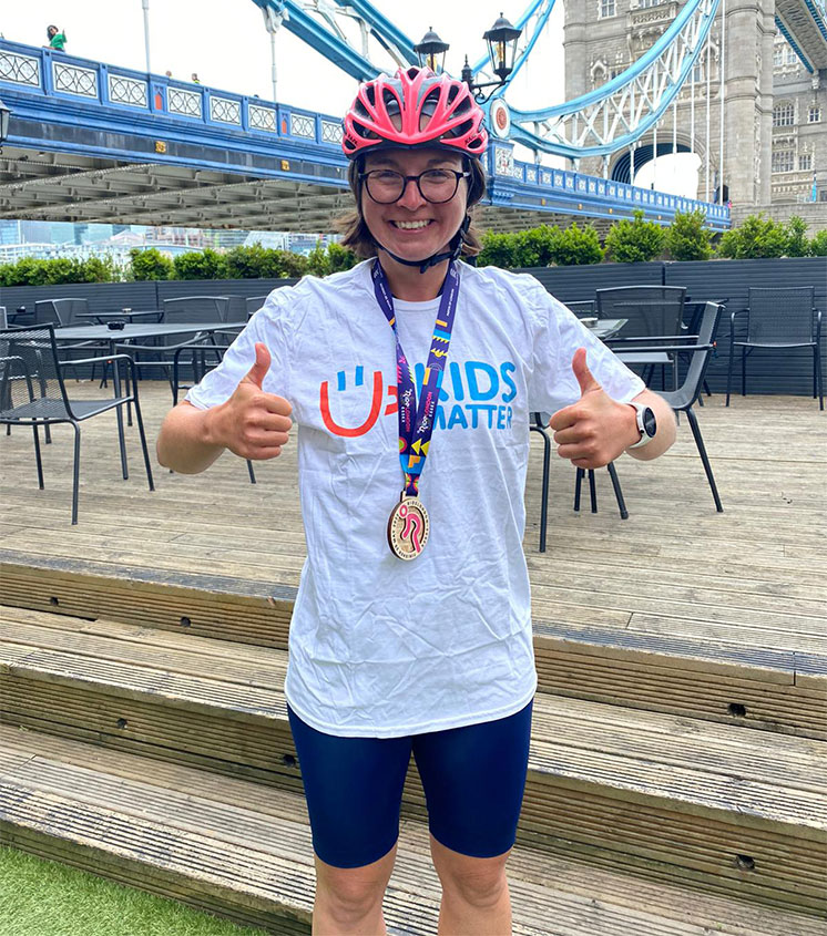 smiling female cyclist wearing a medal and Kids Matter T-shirt standing in front of Tower Bridge