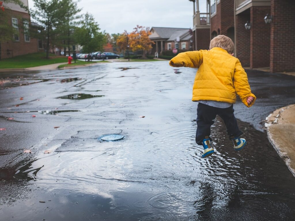 little boy in yellow jacket and wellington boots jumping in puddles in an urban street